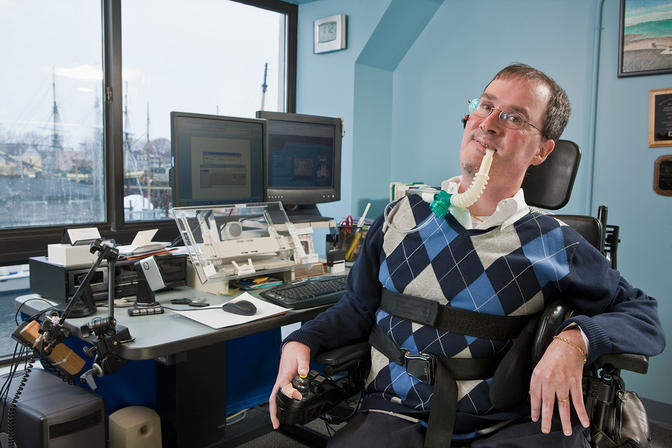 A photo of a man supported in a wheel chair looking at you at his computer in his office at work.