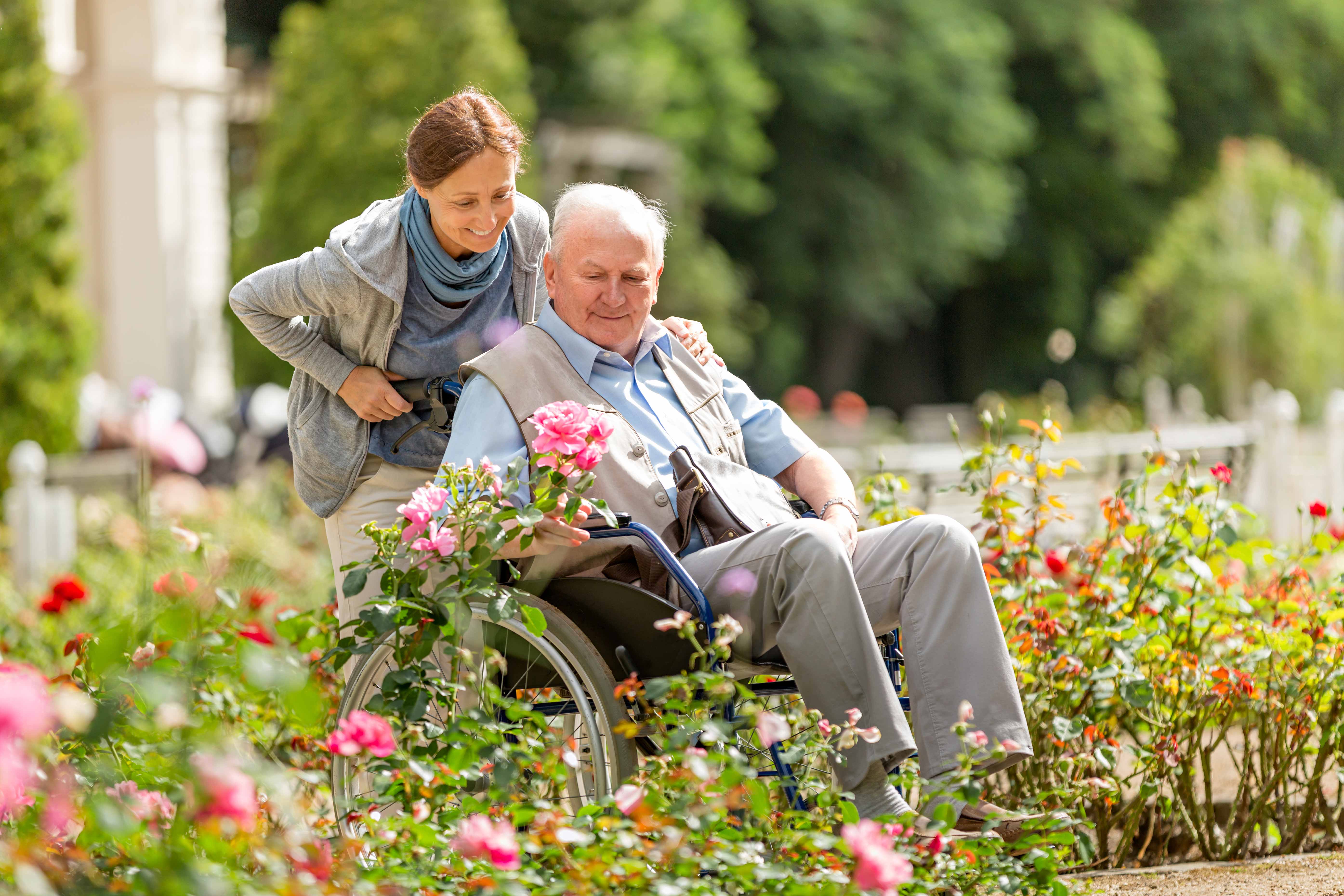 Caregiver and senior man on a wheelchair walking outdoors in a park.