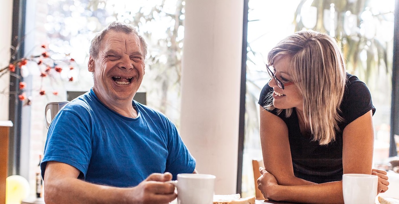 A woman and a man with Down's syndrome sit drinking coffee and laughing at a table