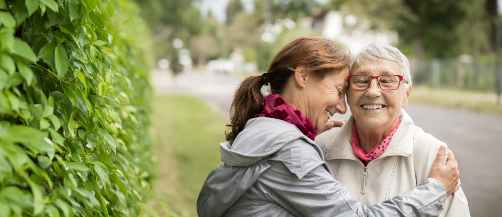 Elderly woman is hugged by her daughter. They're smiling and outside.
